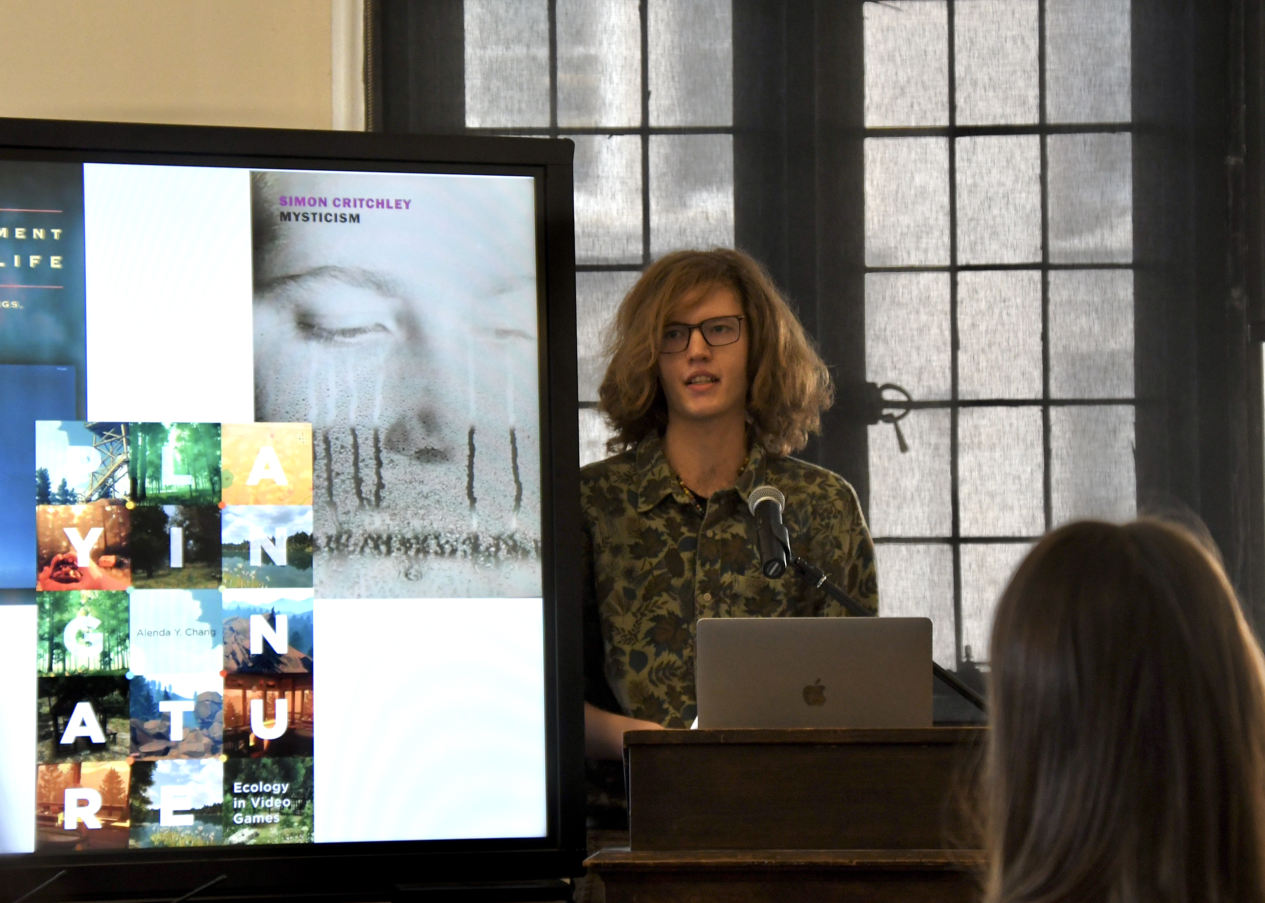 ethan, a white man with long blond hair, stands behind a podium with book covers appearing on the screen to his left